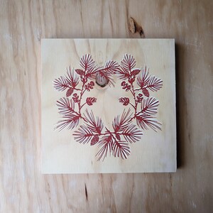 Puede incluir: Placa de madera cuadrada con un diseño de corona de pino roja. La corona presenta ilustraciones detalladas de agujas y piñas de pino. La madera tiene un acabado natural de color claro, el diseño está centrado.