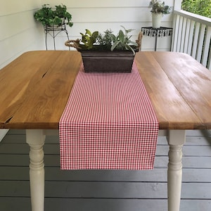 May include: Red and white gingham table runner on a wooden table with white legs. The table is on a gray wooden porch floor. A potted plant is on the table.
