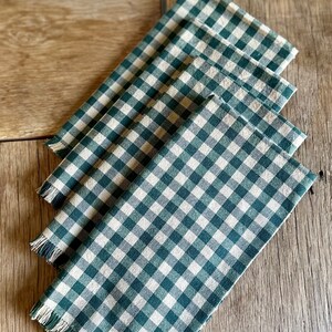 May include: Four green and white gingham napkins with frayed edges. The napkins are folded and stacked on a wooden surface.