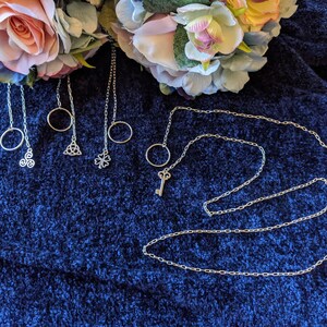 May include: Five silver necklaces with different charms, including a circle, a clover, a Celtic knot, and a key. The necklaces are displayed on a blue fluffy surface.