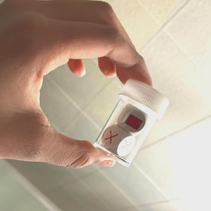 May include: A clear plastic container with a white lid holding three small, white dice. Two dice have a red X on them, and one has a red rectangle. The container is held in a hand.