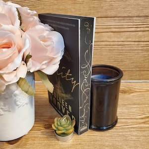 May include: A black book with gold lettering on the spine that reads "Verity" by Colleen Hoover. The book is propped up against a black glass candle jar and a small green succulent candle.