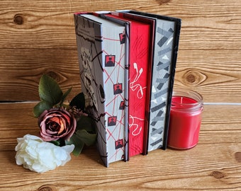 May include: Three hardcover books stacked on a wooden surface. The book on the bottom has a black and gray geometric pattern. The middle book is red with white text. The top book has a gray and white pattern with red lines and red figures.