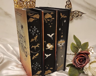 May include: A stack of three hardcover books with black spines and gold illustrations of bats, clouds, and stars. The book on the left has the title "Fourth Wing" and a gold crown sits atop the books. A gold butterfly and flowers are in the background.