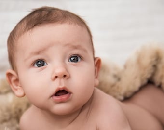 May include: A close-up of a baby's face with big, curious eyes. The baby is lying on a soft, furry blanket.