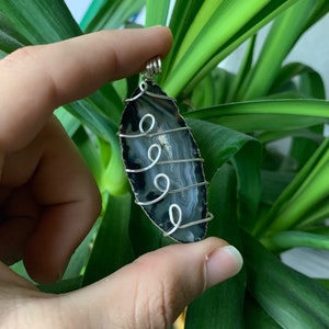 Op de afbeelding: Een zwarte en grijze agaat hanger met zilverdraad. De hanger heeft een druppelvorm en wordt door een hand voor groen gebladerte gehouden. De draad is in decoratieve krullen gewikkeld.