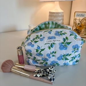 Puede incluir: Un neceser blanco con un estampado floral azul y un interior de cuadros verdes. La bolsa está abierta, revelando el interior. Pinceles de maquillaje, brillo de labios y una pinza para el pelo están junto a la bolsa. La bolsa tiene el texto "meagan leigh design".