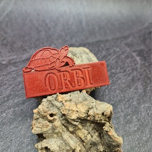 May include: A red 3D printed name tag with a turtle design and the word "ORBI" printed on it.