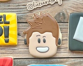 May include: A decorated sugar cookie with a cartoon image of a Roblox character with brown hair and a white smile. The cookie is decorated with brown, white, and black icing.