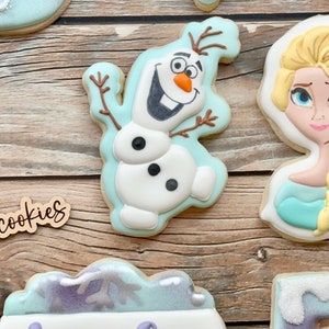 May include: A set of six decorated sugar cookies featuring a snowflake, Olaf from Frozen, Elsa from Frozen, a cookie with the name Amelia, and two cookies with a purple and blue snowflake design.