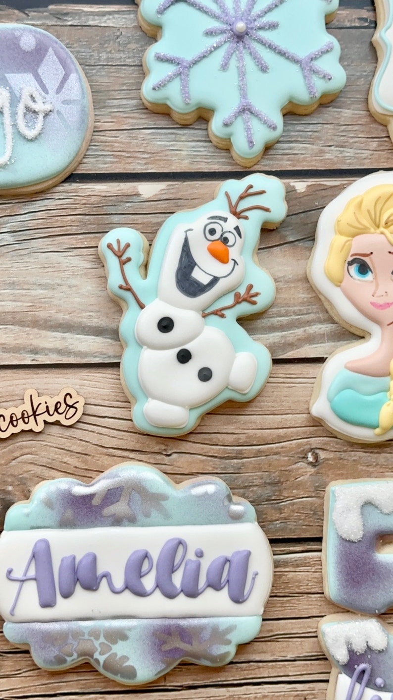 May include: A set of six decorated sugar cookies featuring a snowflake, Olaf from Frozen, Elsa from Frozen, a cookie with the name Amelia, and two cookies with a purple and blue snowflake design.