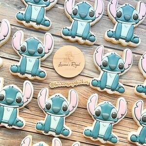 Peut inclure: Une collection de 11 biscuits en sucre d&eacute;cor&eacute;s en bleu et blanc repr&eacute;sentant un personnage de dessin anim&eacute; avec de grands yeux et des oreilles roses. Les biscuits sont d&eacute;cor&eacute;s de gla&ccedil;age bleu, blanc et rose. Les biscuits sont dispos&eacute;s sur une surface en bois. Le texte "Luciana's Royal Cookies" est visible sur un cercle en bois.