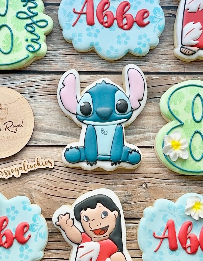 Peut inclure: Un ensemble de biscuits en sucre d&eacute;cor&eacute;s repr&eacute;sentant un personnage bleu Stitch de Lilo & Stitch, un biscuit vert avec le chiffre 8 et deux biscuits avec le nom "Abbe" &eacute;crit en rouge. Les biscuits sont d&eacute;cor&eacute;s de motifs floraux bleus et blancs.