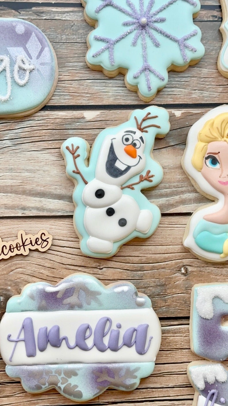 May include: A set of six decorated sugar cookies featuring a snowflake, Olaf from Frozen, Elsa from Frozen, a cookie with the word "cookies" on it, a cookie with the name "Amelia" on it, and a cookie with a snowflake design.