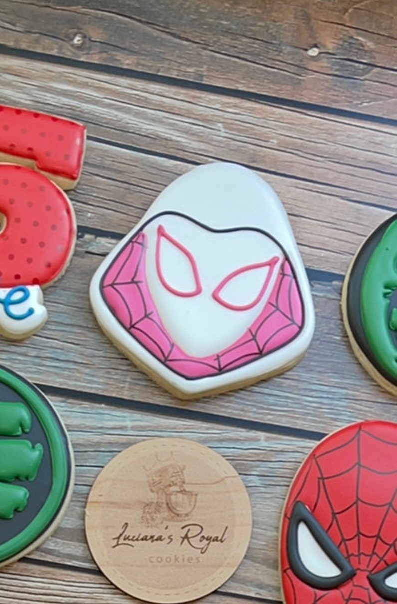 May include: A set of decorated sugar cookies featuring superhero characters. The cookies are decorated with white, red, black, and green icing. The cookies are arranged on a wooden surface. The cookies are decorated with the text "Luciana's Royal Cookies".