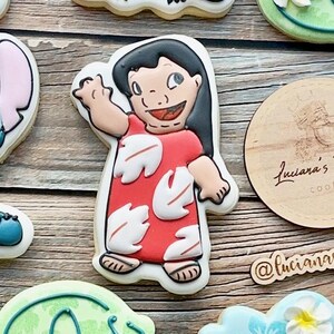 May include: A set of decorated sugar cookies featuring characters from the Disney movie Lilo & Stitch. The cookies are decorated with colourful icing and feature Lilo, Stitch, and a flower.