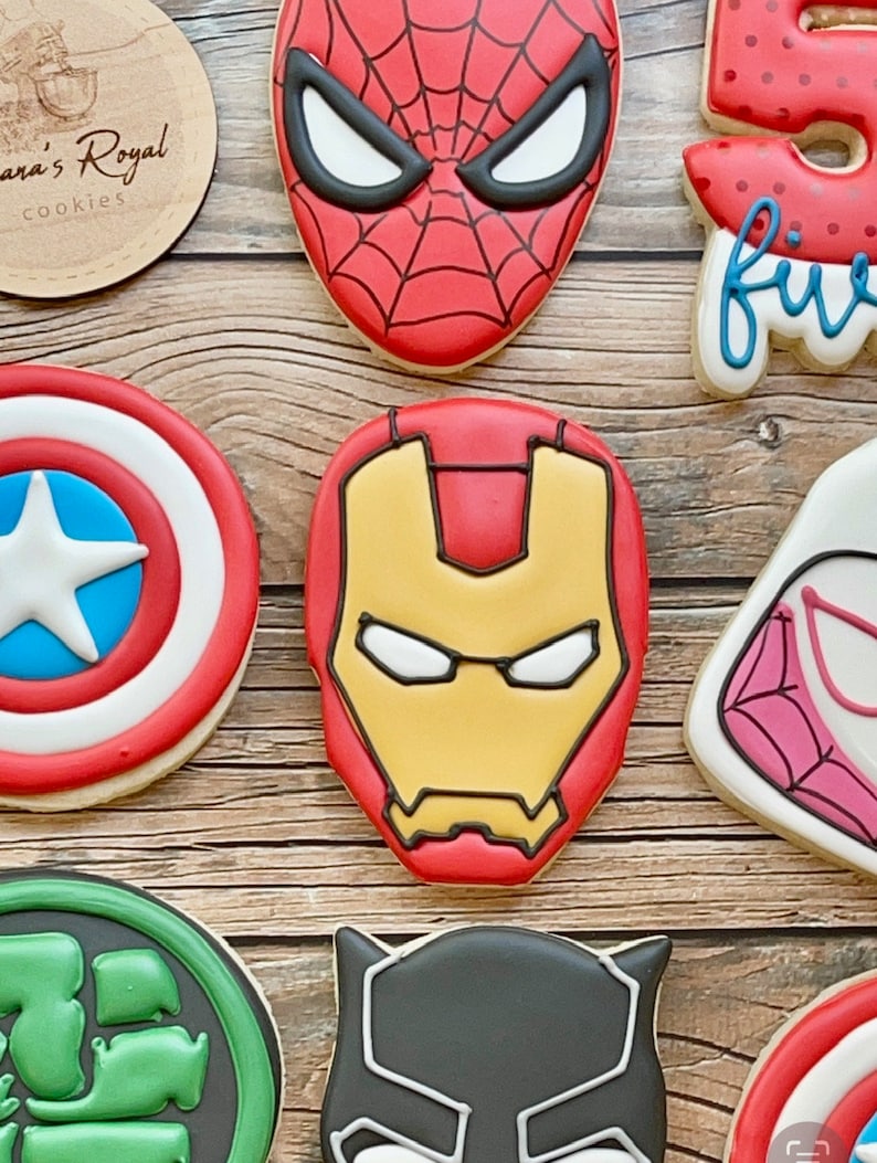 May include: A collection of superhero-themed cookies, including Spider-Man, Iron Man, Captain America, Hulk, Black Panther, and a red and white cookie with the word "five" on it.