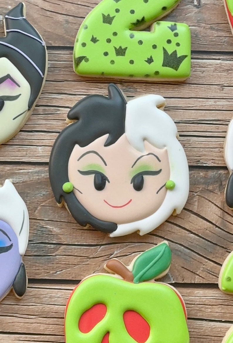 May include: A set of four cookies decorated with characters from the Disney film "101 Dalmatians". The cookies feature a green and black cookie with a crown design, a cookie with a close-up of Cruella de Vil's face, a purple cookie with a side profile of Cruella de Vil, and a green apple cookie with a red skull design.