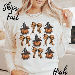 May include: An ivory long-sleeve shirt featuring a repeating graphic pattern of orange pumpkins wearing black witch hats, interspersed with brown ribbons, small bats, and star icons. The design is centered on the front of the garment.