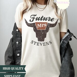 May include: A beige t-shirt with a distressed brown bull skull graphic. The text "Future Mrs. Stevens" is printed on the shirt.