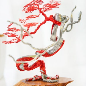 Op de afbeelding: Een sculptuur van een bonsaiboom van rood en zilverdraad op een houten voet. De boom heeft een gedraaide stam en takken met kleine, rode bladeren.