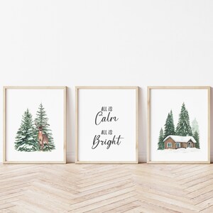 Puede incluir: Tres impresiones enmarcadas con un tema invernal. La impresión de la izquierda presenta un ciervo de pie en un bosque nevado. La impresión del medio tiene el texto "All is Calm, All is Bright". La impresión de la derecha muestra una pequeña cabaña en un bosque nevado.