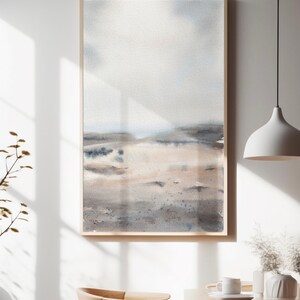 Peut inclure: Une peinture &agrave; l'aquarelle encadr&eacute;e d'un paysage avec un ciel bleu clair, des collines brunes et grises et une plage de sable blanc. La peinture est dans un cadre en bois et est accroch&eacute;e &agrave; un mur blanc.
