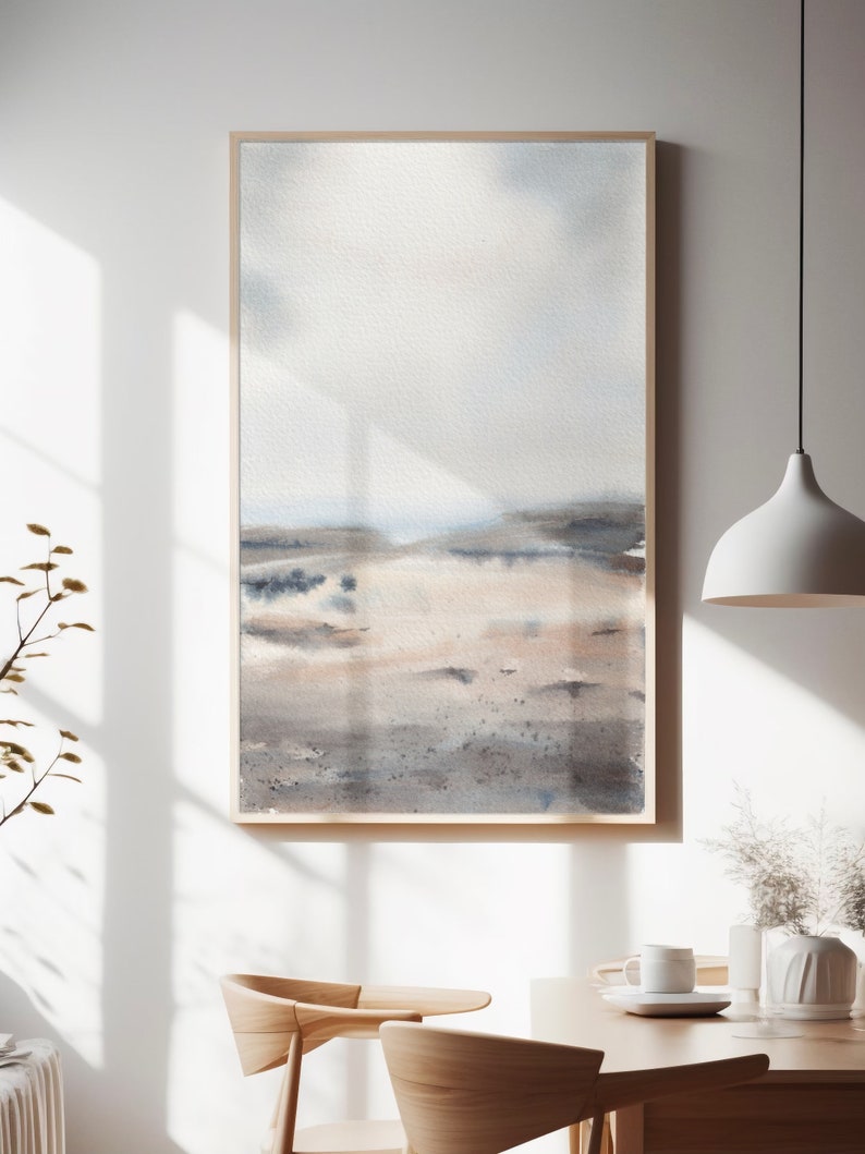 Peut inclure: Une peinture &agrave; l'aquarelle encadr&eacute;e d'un paysage avec un ciel bleu clair, des collines brunes et grises et une plage de sable blanc. La peinture est dans un cadre en bois et est accroch&eacute;e &agrave; un mur blanc.