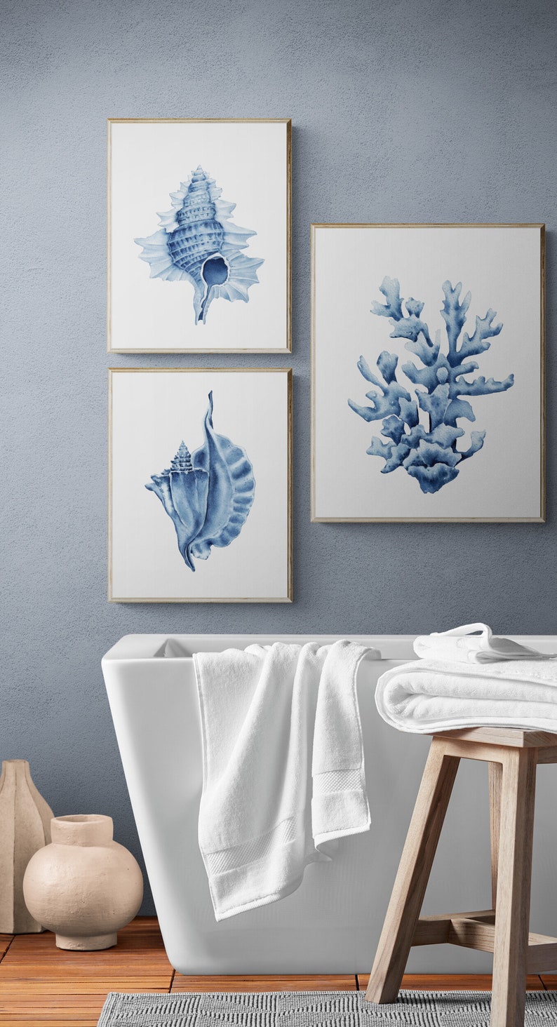 May include: Three framed watercolor prints of seashells and coral in shades of blue on a white background. The prints are hanging on a light blue wall above a white bathtub and a wooden stool with white towels.