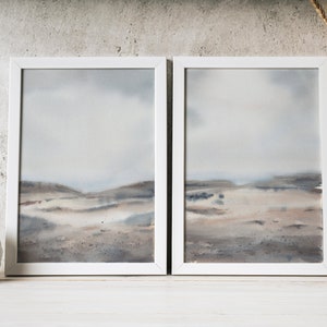 Peut inclure: Deux aquarelles encadr&eacute;es d'un paysage avec une palette de couleurs sourdes de gris, de bruns et de bleus. Les peintures sont accroch&eacute;es &agrave; un mur blanc avec un vase blanc de tiges de bl&eacute; s&eacute;ch&eacute;es &agrave; droite.