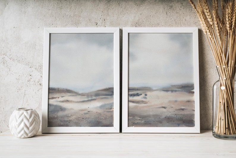 Peut inclure: Deux aquarelles encadr&eacute;es d'un paysage avec une palette de couleurs sourdes de gris, de bruns et de bleus. Les peintures sont accroch&eacute;es &agrave; un mur blanc avec un vase blanc de tiges de bl&eacute; s&eacute;ch&eacute;es &agrave; droite.