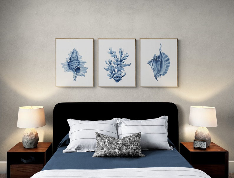 May include: Three framed watercolor prints of blue seashells and coral on a white wall above a black headboard with white and blue striped pillows and a gray and white patterned throw pillow on a bed with a blue duvet and white sheets.