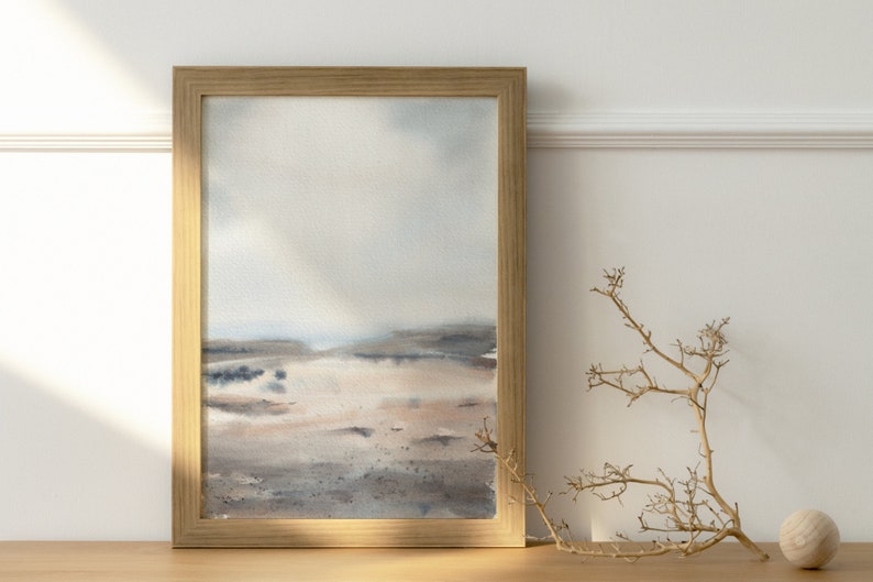 Peut inclure: Une peinture &agrave; l'aquarelle encadr&eacute;e d'un paysage avec un ciel bleu clair, une terre brune et grise et une touche d'eau au loin. La peinture est dans un cadre en bois brun clair.