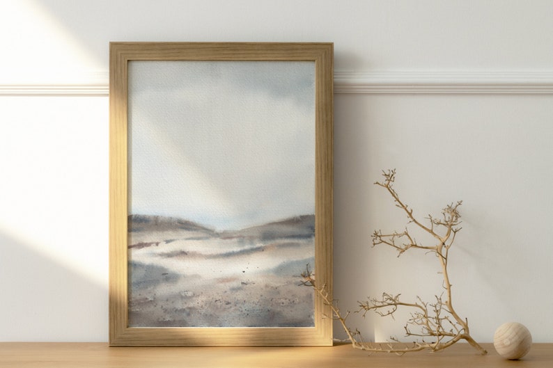 Peut inclure: Une peinture &agrave; l'aquarelle encadr&eacute;e d'un paysage avec un ciel bleu clair et des collines brunes. La peinture est dans un cadre en bois et est accroch&eacute;e &agrave; un mur blanc.
