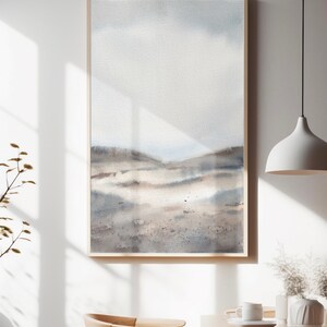 Peut inclure: Une peinture &agrave; l'aquarelle encadr&eacute;e d'un paysage avec un ciel bleu clair et des collines brunes. La peinture est dans un cadre en bois clair et est accroch&eacute;e &agrave; un mur blanc.