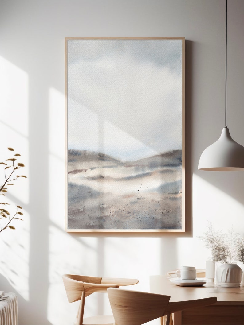 Peut inclure: Une peinture &agrave; l'aquarelle encadr&eacute;e d'un paysage avec un ciel bleu clair et des collines brunes. La peinture est dans un cadre en bois clair et est accroch&eacute;e &agrave; un mur blanc.