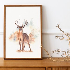 May include: A watercolor painting of a white-tailed deer standing in a forest. The deer has large antlers and is looking directly at the viewer. The background is a soft watercolor wash of brown and green.