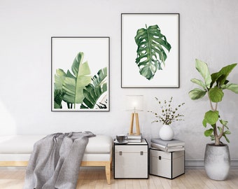 May include: Two framed botanical prints of green leaves, one with a single large leaf and the other with multiple smaller leaves, hanging on a white wall above a white sofa with a grey throw blanket. A small wooden table with a lamp, a cup, and books sits next to the sofa. A potted plant with large green leaves is on the right side of the image.