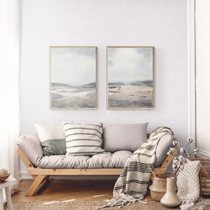 Peut inclure: Un canap&eacute; gris clair avec deux coussins et une couverture. Le canap&eacute; est devant un mur blanc avec deux tableaux de paysages encadr&eacute;s. Les peintures sont dans un sch&eacute;ma de couleurs gris clair avec une touche de bleu. Il y a un vase avec une branche de coton blanche et un panier avec une couverture en tricot blanc sur le sol.