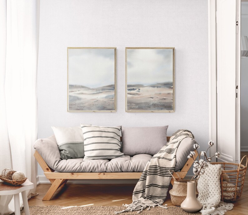 Peut inclure: Un canap&eacute; gris clair avec deux coussins et une couverture. Le canap&eacute; est devant un mur blanc avec deux tableaux de paysages encadr&eacute;s. Les peintures sont dans un sch&eacute;ma de couleurs gris clair avec une touche de bleu. Il y a un vase avec une branche de coton blanche et un panier avec une couverture en tricot blanc sur le sol.