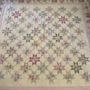 Pode incluir: Um edredom grande de cor creme com um design de patchwork que apresenta um padrão repetitivo de estrelas multicoloridas. O edredom tem uma borda rosa e branca.