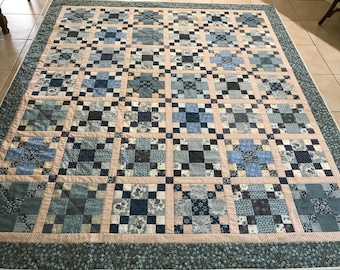 May include: A blue and white quilt with a patchwork design. The quilt is made up of squares of different fabrics, all in shades of blue and white. The quilt is edged with a dark blue border.
