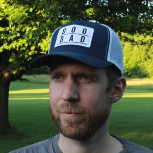 May include: A man wearing a blue and white trucker hat with a white patch that says "DOG DAD" in black letters. He is wearing a grey shirt and looking to the right.