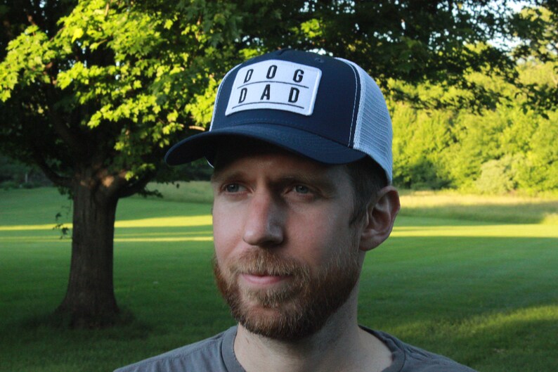 May include: A man wearing a blue and white trucker hat with a white patch that says "DOG DAD" in black letters. He is wearing a grey shirt and looking to the right.