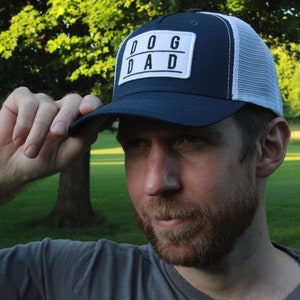 May include: A navy blue and white trucker hat with a white patch that says "DOG DAD" in black letters.