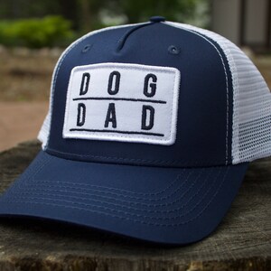 May include: A navy blue trucker hat with a white mesh back. The hat has a white patch on the front that says "DOG DAD" in black block letters.
