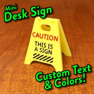 May include: A yellow miniature A-frame desk sign featuring a cutout handle at the top. The sign displays the red text CAUTION above the black text THIS IS A SIGN, followed by a series of black diagonal stripes at the bottom of the front panel.