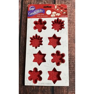 May include: A red silicone candy mold by Wilton featuring ten cavities in various snowflake and star shapes. The packaging displays the text Candy Melts and Candy Mold, with a white background decorated with light gray snowflake patterns.