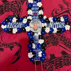 Könnte beinhalten: Ein kreuzförmiges Gedenkarrangement mit blauen, schwarzen und weißen floralen Akzenten. Das Kreuz trägt die Namen "Reggie" und "Nettles" sowie den Text "Your wings were ready but our hearts were not". Ein Foto befindet sich in der Mitte.
