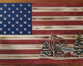 May include: A wooden American flag with a distressed finish. The flag features a carved design of a deer standing in a forest with trees. The flag is painted in red, white, and blue.